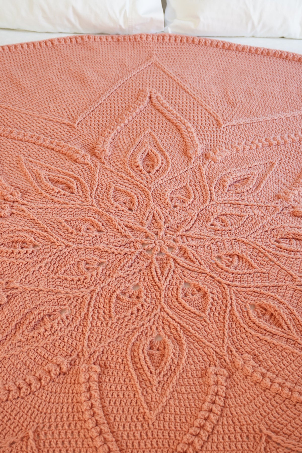 A peach-pink crocheted blanket with an intricate mandala pattern, spread out on a bed with white pillows visible at the top.