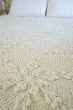 A close-up of a cream-colored crocheted blanket with intricate textured patterns and snowflake motifs, styled on a bed with white pillows in the background.