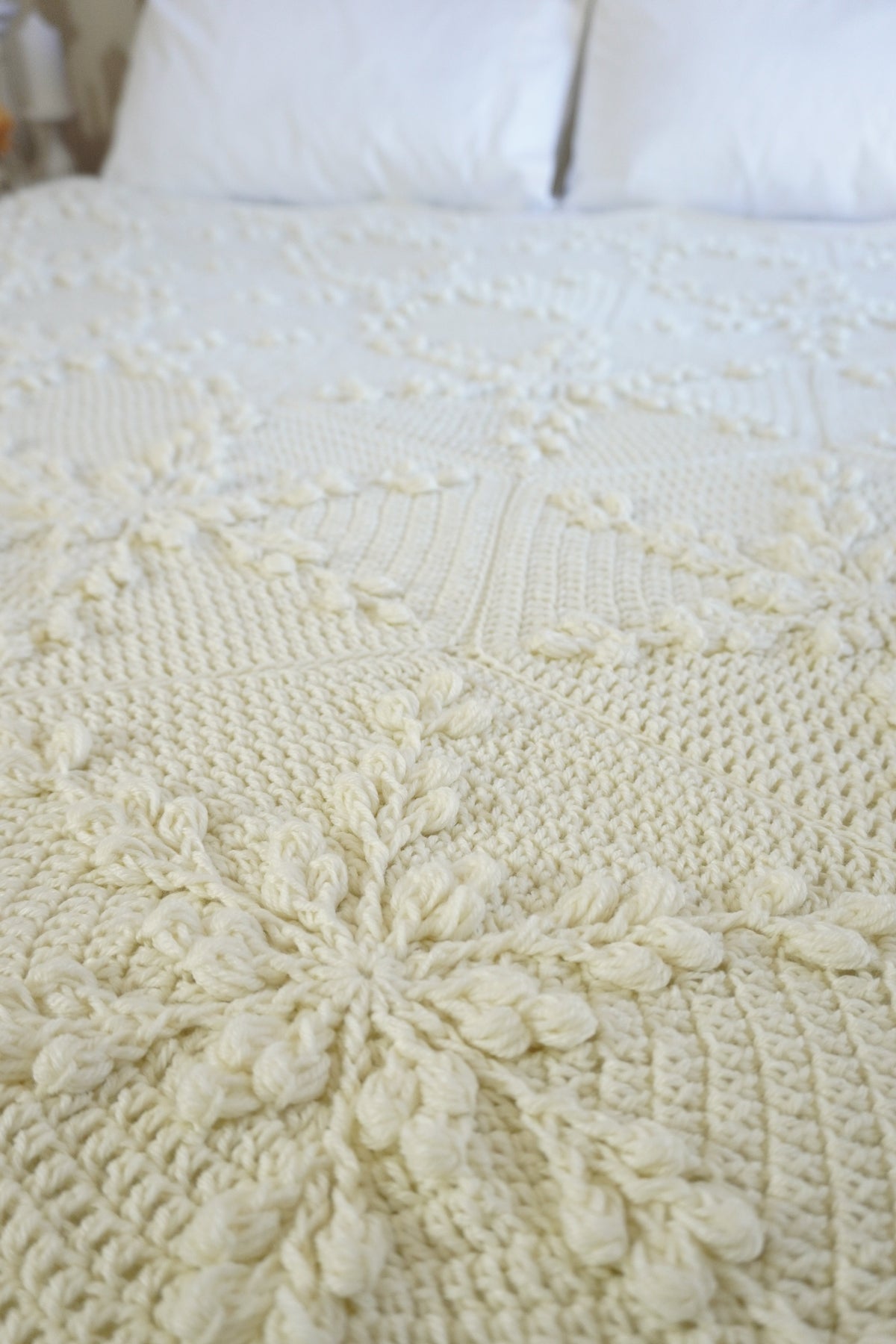 A close-up of a cream-colored crocheted blanket with intricate textured patterns and snowflake motifs, styled on a bed with white pillows in the background.
