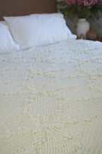 A close-up of a cream-colored crocheted blanket with snowflake design, styled on a bed with white pillows and a blurred background showing a vase of pink flowers.