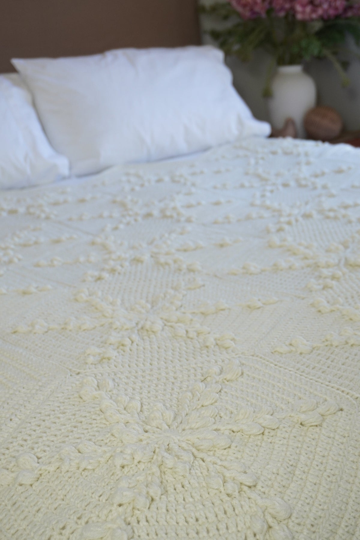 A close-up of a cream-colored crocheted blanket with snowflake design, styled on a bed with white pillows and a blurred background showing a vase of pink flowers.