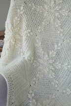 A close-up of a cream-colored crocheted blanket with textured snowflake pattern draped over a chair.