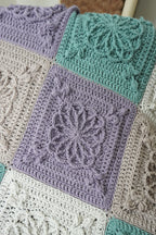 A close-up of a handmade crocheted patchwork blanket featuring textured square panels in lavender, mint green, and cream.