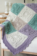 A crocheted patchwork throw blanket draped over a light wood chair in a softly styled room. The blanket features alternating textured square panels in lavender, mint green, and cream, finished with a scalloped lavender border.