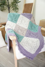 A handmade crochet blanket with patchwork granny squares in lavender, mint green, and cream, draped over a light wood chair in a cozy room setting.