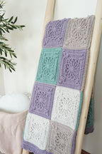 A handmade crocheted patchwork blanket with lavender, mint green, and white granny squares draped over a wooden ladder in a bright room.