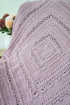 A close-up of a handmade crochet blanket in a dusty lavender-purple color, featuring intricate openwork lace patterns and concentric square designs. The blanket is draped over a chair.
