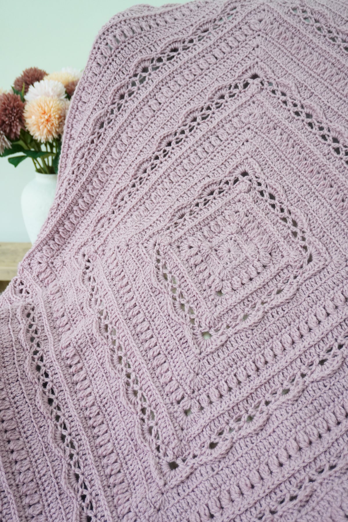 A close-up of a handmade crochet blanket in a dusty lavender-purple color, featuring intricate openwork lace patterns and concentric square designs. The blanket is draped over a chair.