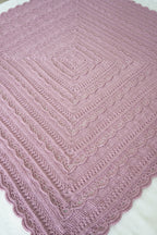 A square pink crocheted blanket with a scalloped edge and textured concentric diamond patterns, laid flat on a white surface.