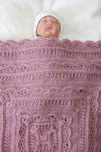A close-up of a sleeping newborn baby wrapped in a dusty pink crocheted blanket with a white hat, lying on a white blanket.