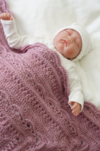 A sleeping newborn baby doll or realistic baby figurine wearing a white outfit and hat, lying on a white blanket under a dusty pink textured crochet blanket.