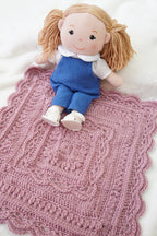 A doll with blonde pigtails sits on a pink crocheted blanket on a white textured fabric background.