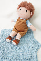 A plush doll with reddish-brown hair, a blue and white striped shirt, brown corduroy overalls, and brown shoes, lying on a light blue crocheted blanket against a white background.