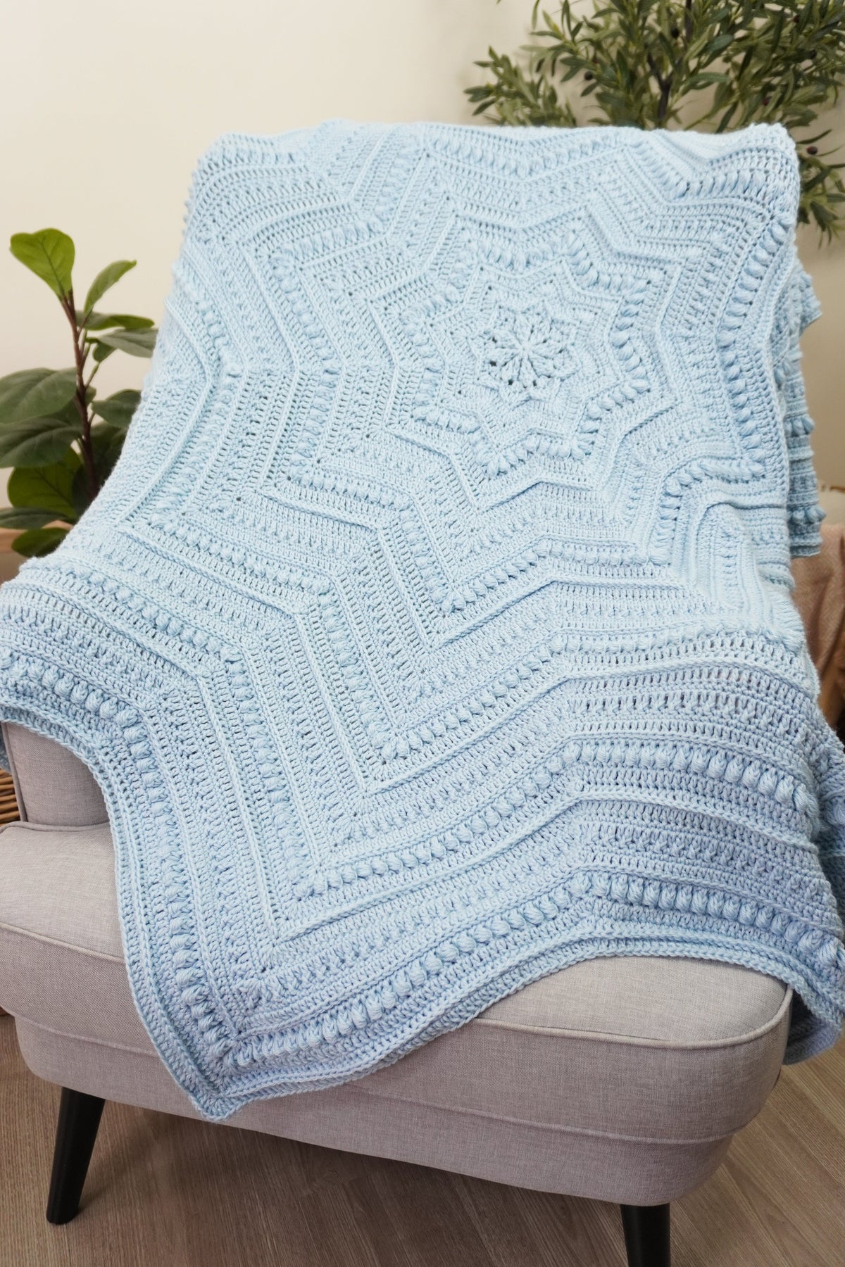 A light blue crocheted blanket with a geometric starburst pattern draped over a light gray upholstered armchair in a styled indoor setting with plants and wood flooring.