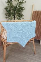 A light blue crocheted blanket draped over a rattan bassinet in a styled indoor room with a potted plant and wood flooring.