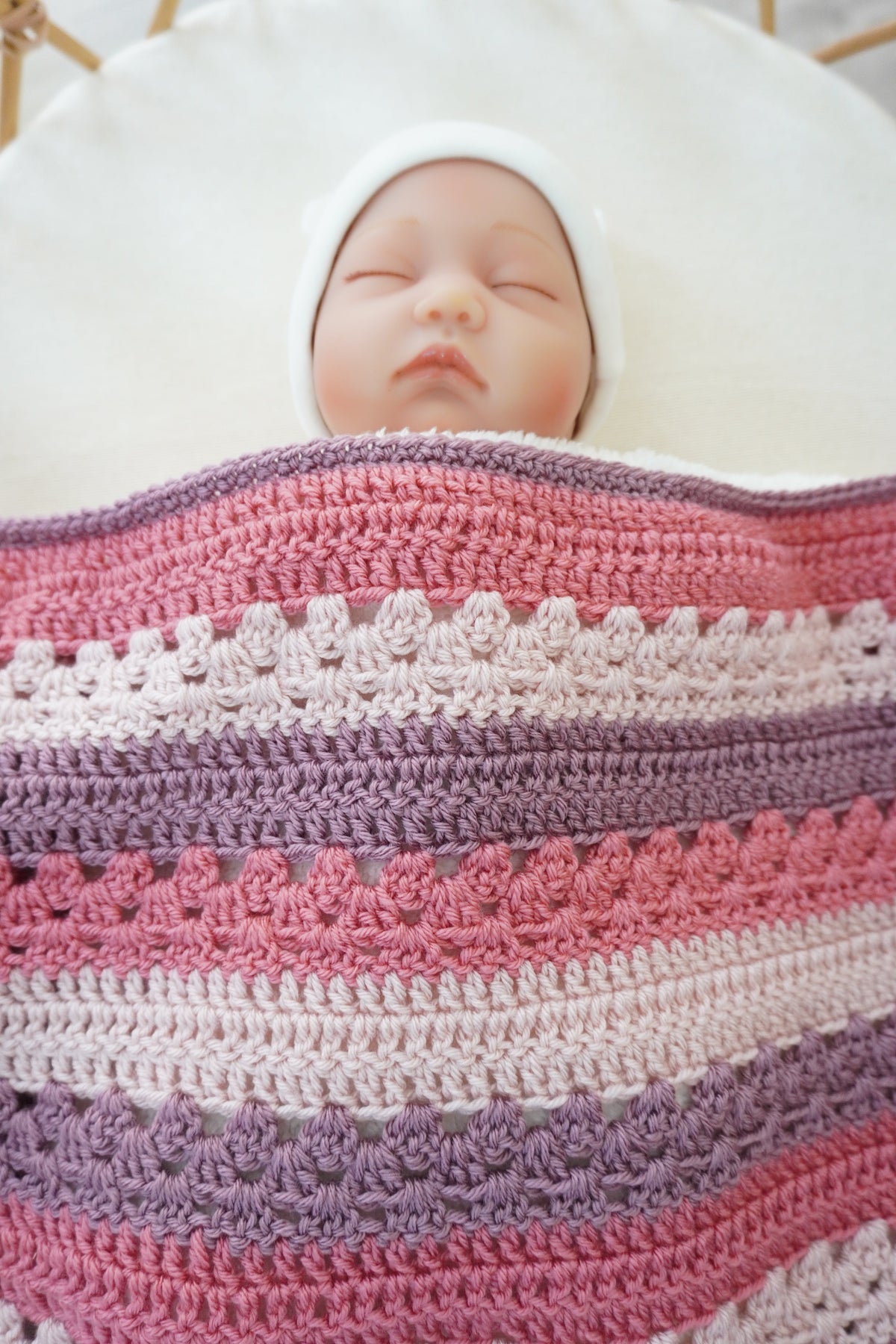 A close-up of a newborn baby doll wrapped in a soft, striped crocheted blanket with pink, mauve, and cream stripes, wearing a white bonnet and lying on a white cushion.