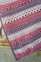 A close-up of a handmade crocheted blanket with horizontal stripes in dusty pink, mauve purple, and cream, draped over a light wood chair.