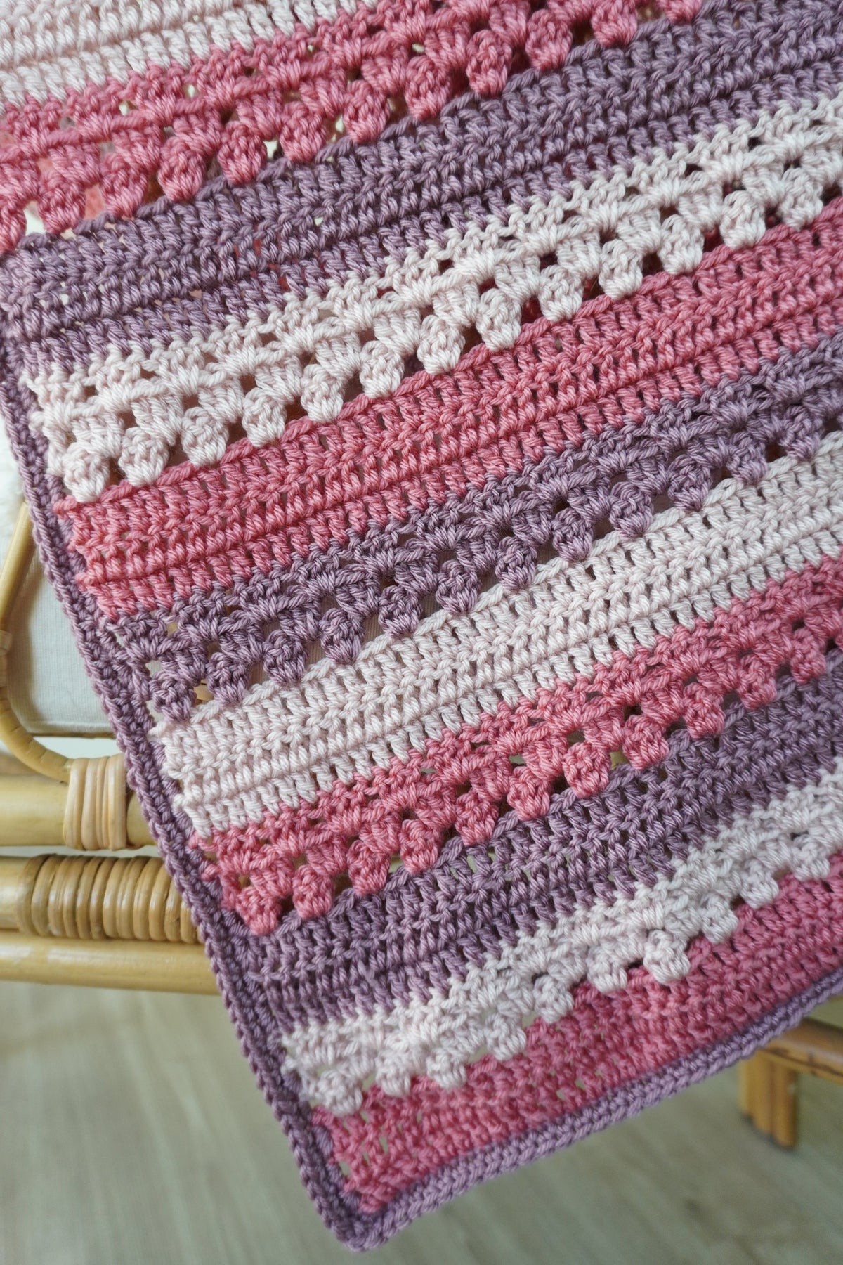 A close-up of a handmade crocheted blanket with horizontal stripes in dusty pink, mauve purple, and cream, draped over a light wood chair.
