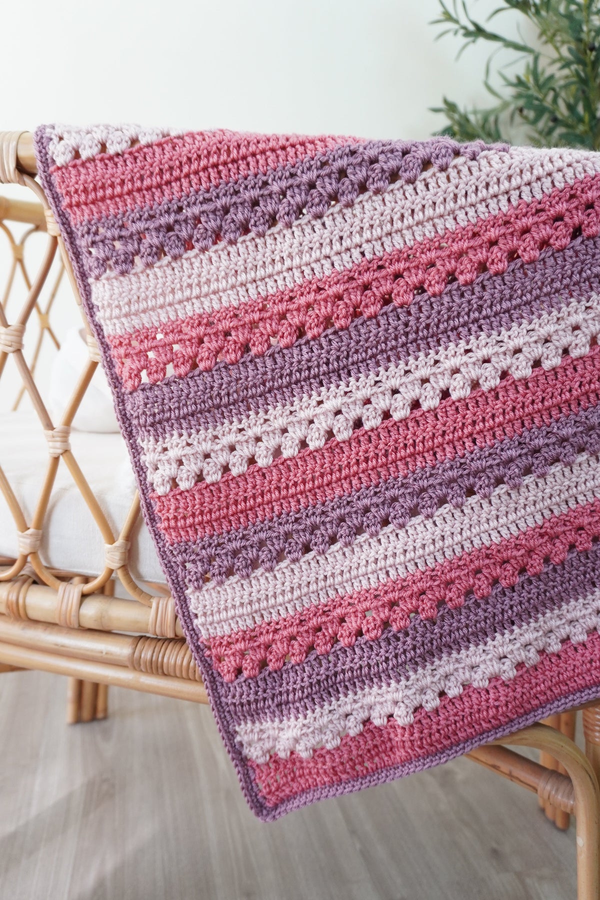 A handmade crocheted blanket with horizontal stripes in shades of pink, mauve, and lavender, draped over a rattan chair in a bright room.