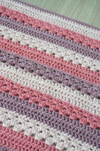 Close-up of a crocheted balanket with horizontal stripes in cream, dusty pink, mauve, and light gray.