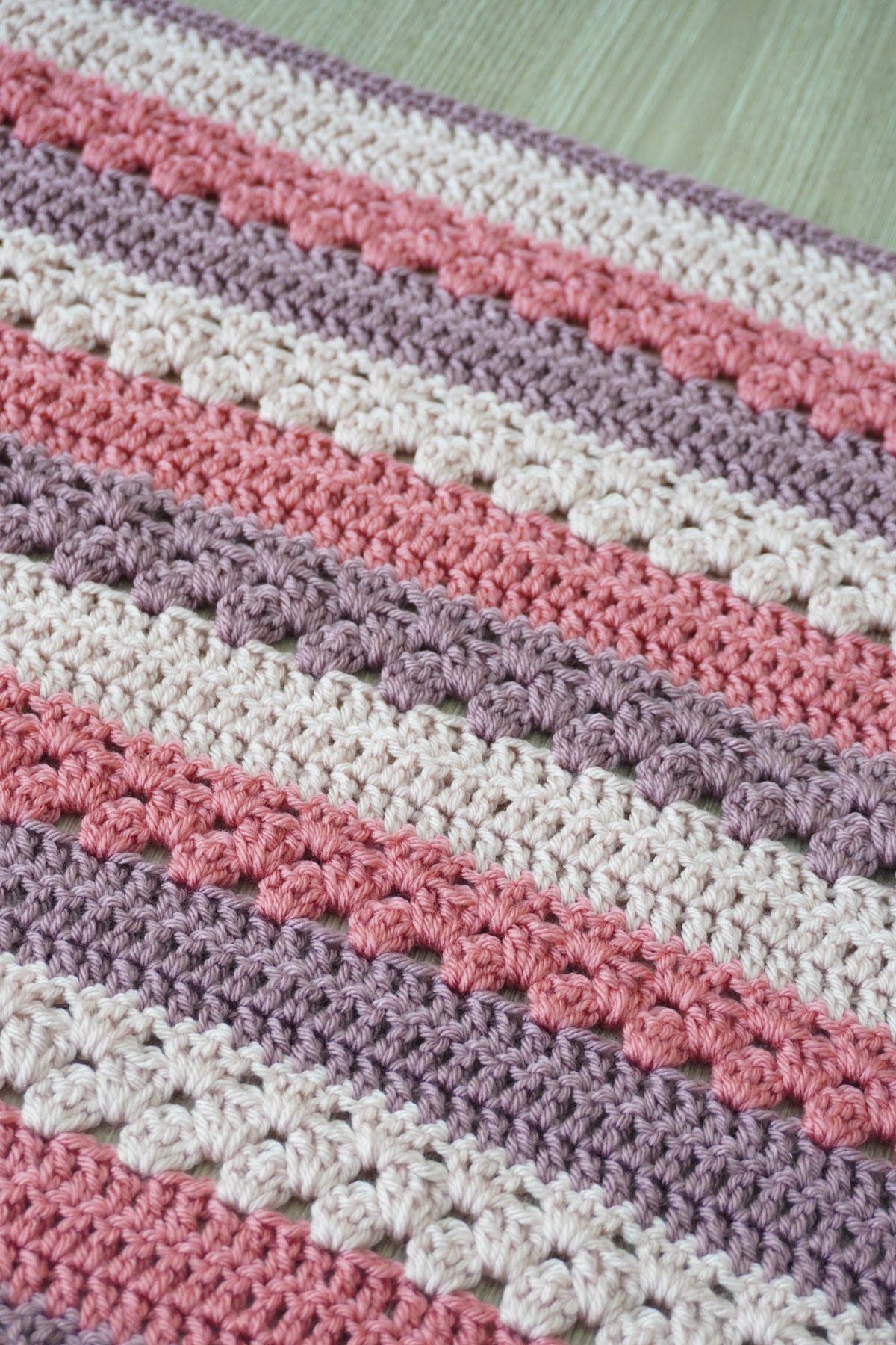 Close-up of a crocheted balanket with horizontal stripes in cream, dusty pink, mauve, and light gray.