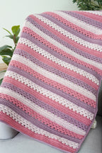 A handmade crocheted baby blanket with horizontal stripes in dusty pink, lavender purple, and cream white, draped over a light gray sofa with green plants in the background.