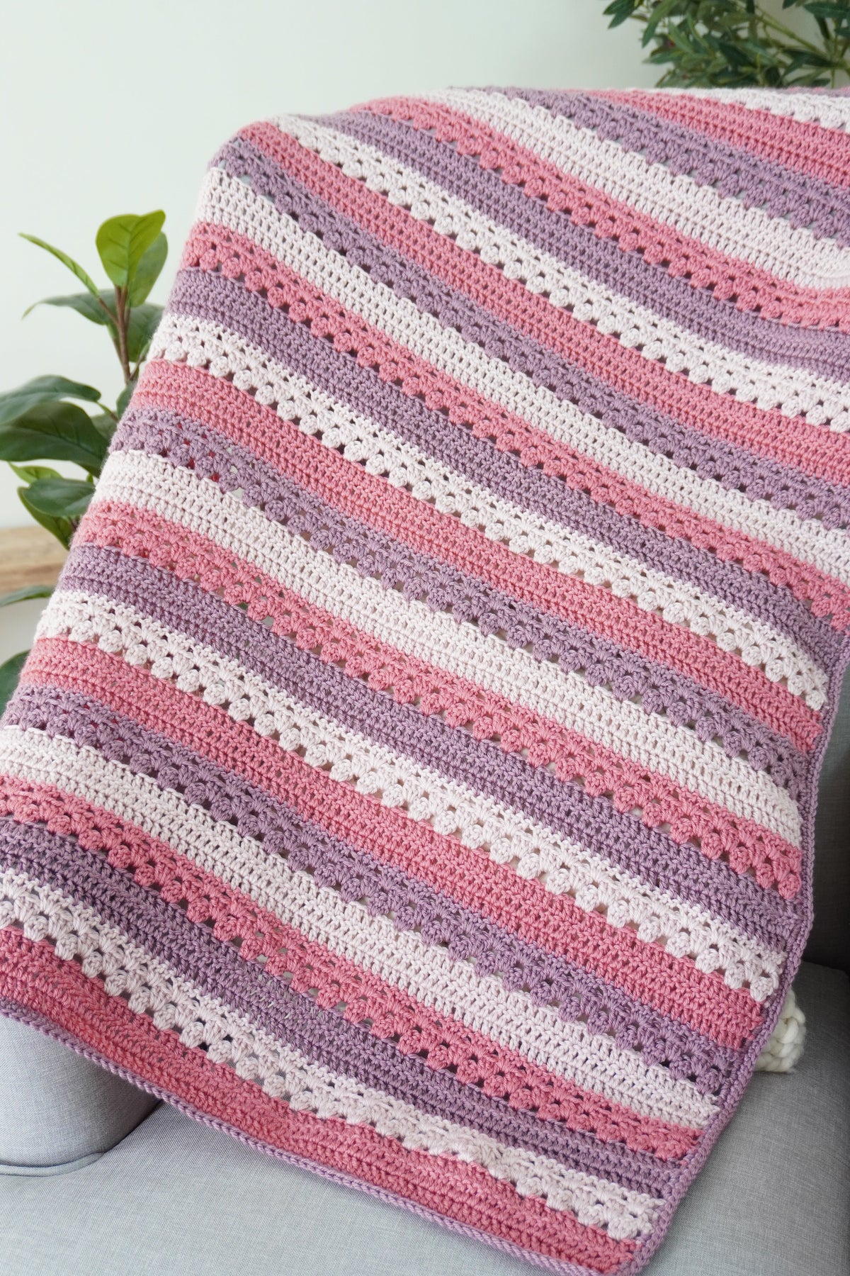 A handmade crocheted baby blanket with horizontal stripes in dusty pink, lavender purple, and cream white, draped over a light gray sofa with green plants in the background.