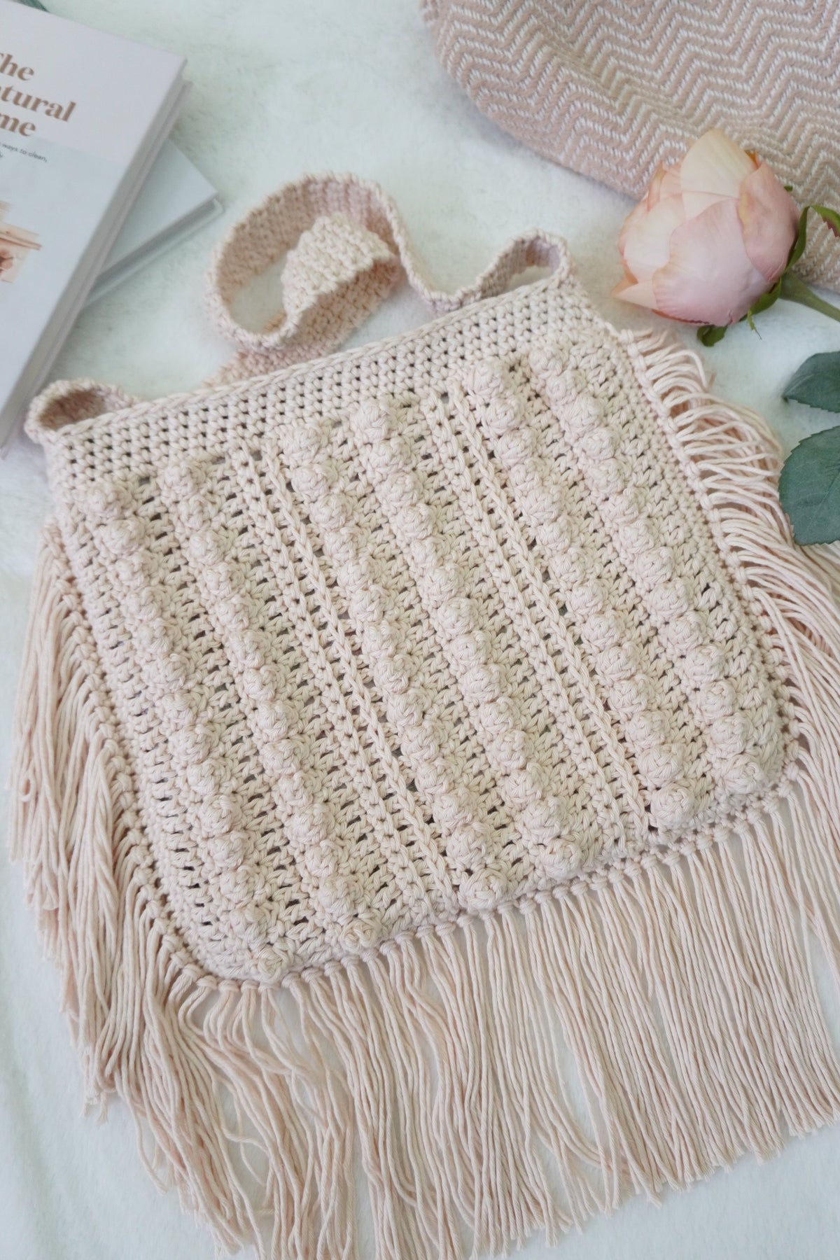 A cream-colored crocheted shoulder bag with fringe detailing, styled on a white textured blanket with a pink rose, a book, and a patterned textile in the background.
