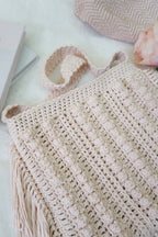 A close-up of a hand-crocheted beige tote bag with textured vertical stripes and fringe, styled on a white surface with a book, a pink rose, and another crocheted textile in the background.