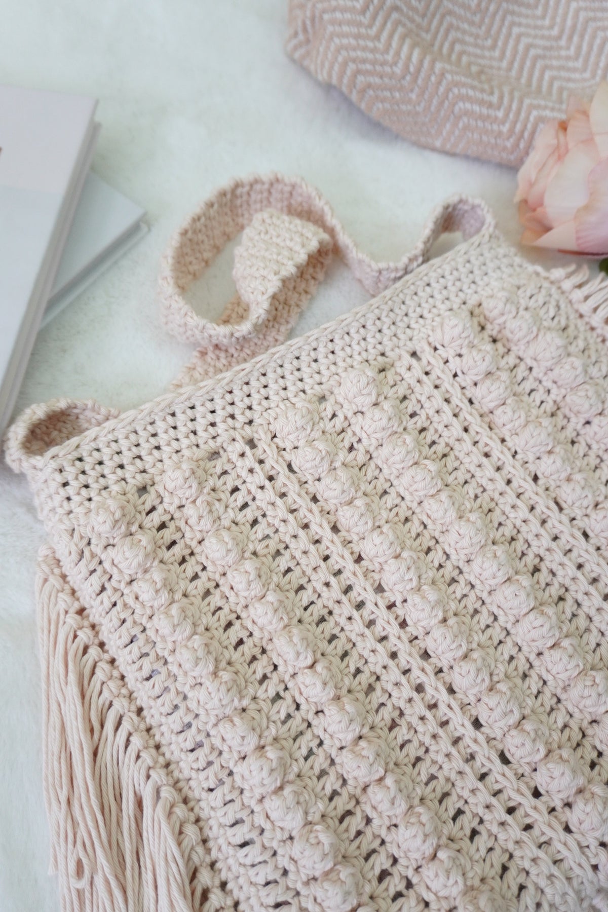 A close-up of a hand-crocheted beige tote bag with textured vertical stripes and fringe, styled on a white surface with a book, a pink rose, and another crocheted textile in the background.