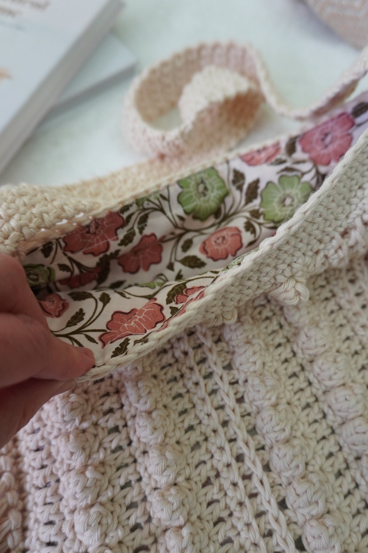 A close-up of a cream-colored crocheted bag with a floral-patterned lining, held open by a hand. The bag features a textured crochet body and a matching cream strap, with a vintage-style book visible in the background.