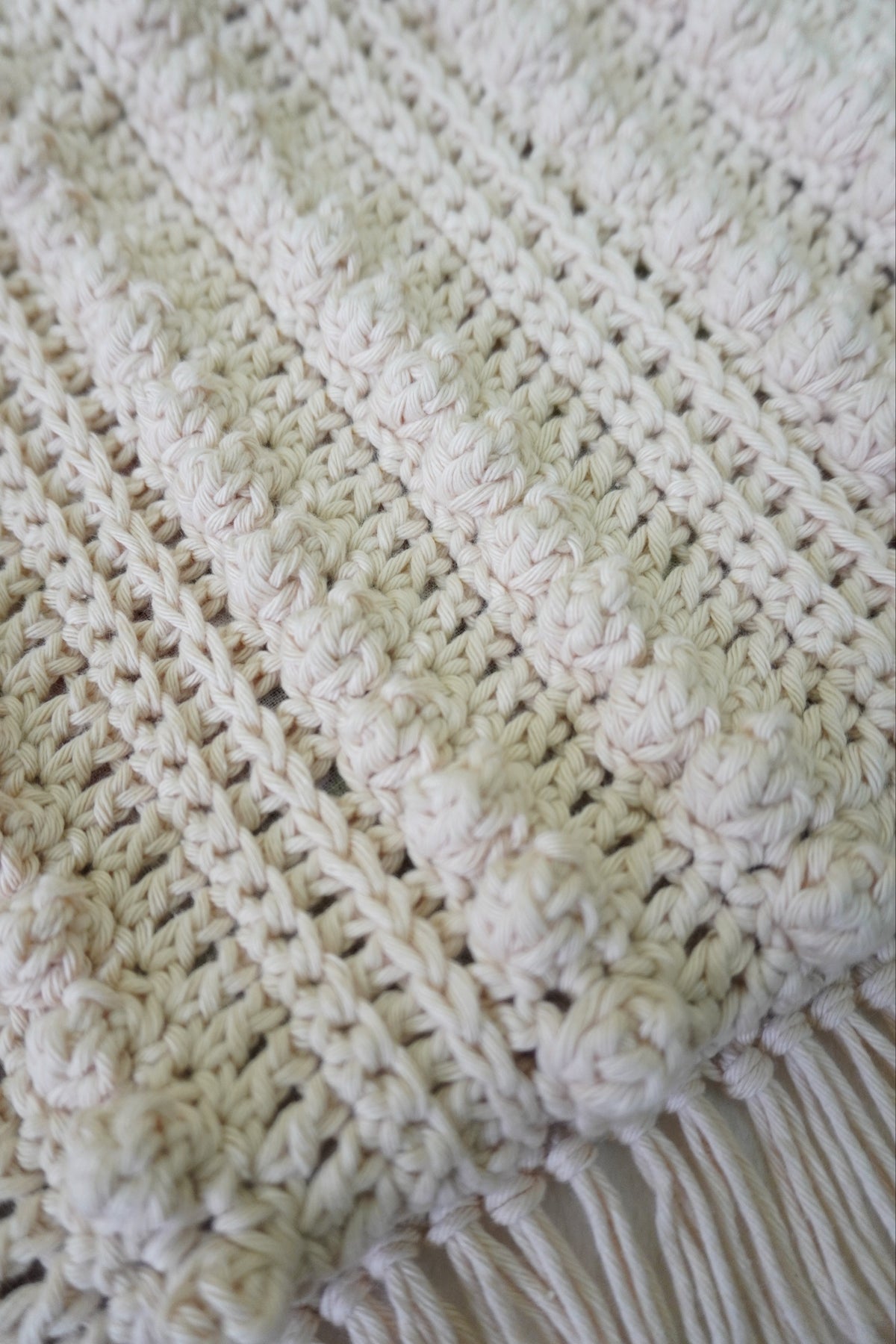 Close-up of a cream-colored crocheted textile with a textured crochet pattern and fringe along the bottom edge.
