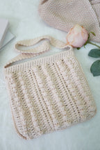 A cream-colored crocheted shoulder bag with a textured vertical rib pattern, styled on a white textured blanket with a pink rose and a book in the background.