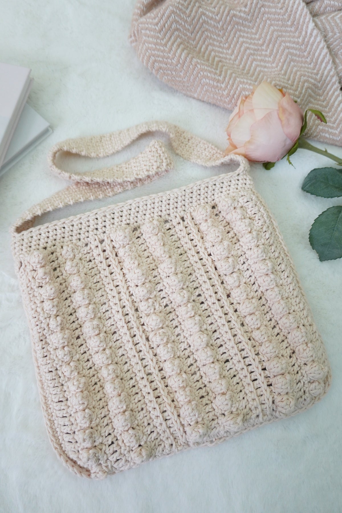 A cream-colored crocheted shoulder bag with a textured vertical rib pattern, styled on a white textured blanket with a pink rose and a book in the background.
