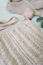 A close-up of a handmade crochet tote bag in a soft oatmeal beige color, styled with a pink rose and a beige chevron scarf on a white fabric background.