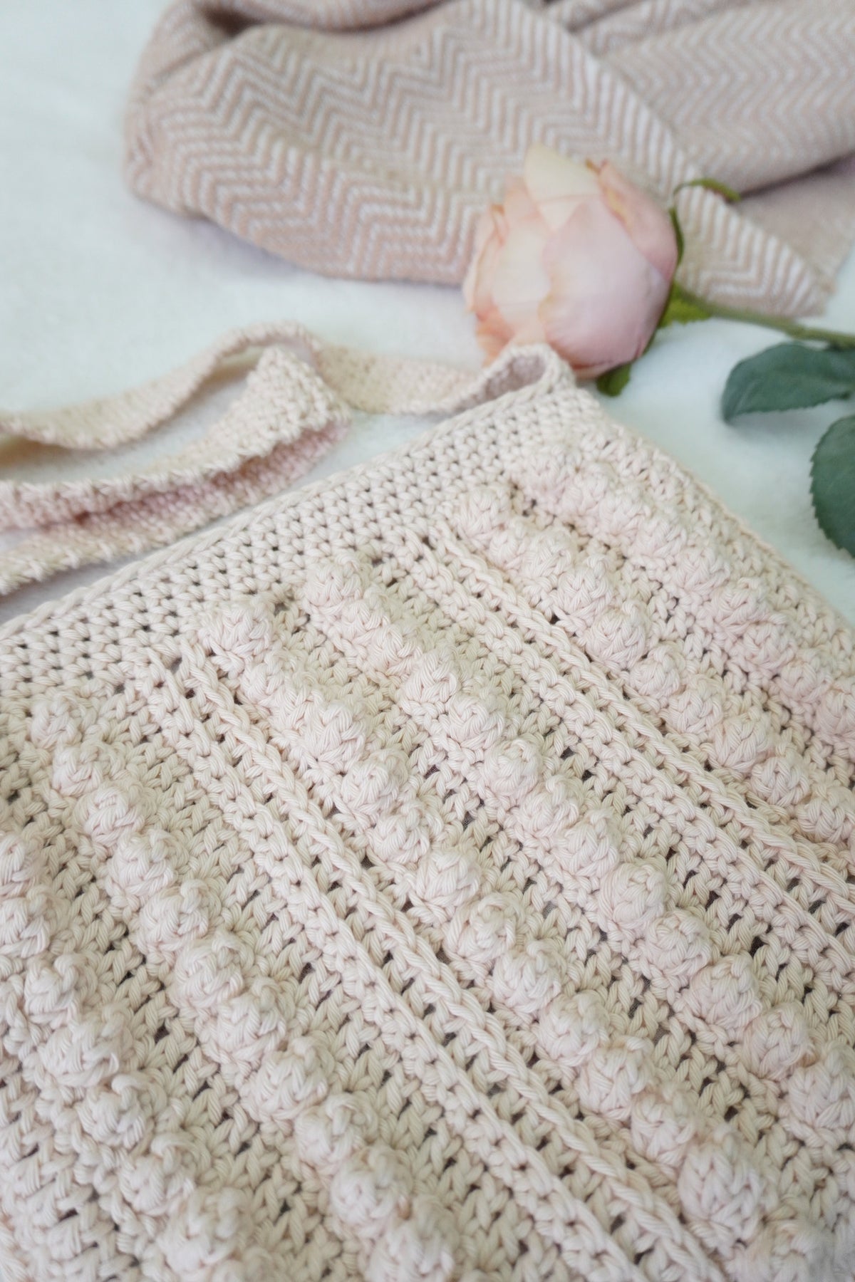 A close-up of a handmade crochet tote bag in a soft oatmeal beige color, styled with a pink rose and a beige chevron scarf on a white fabric background.