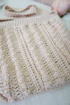 A close-up of a handmade, chunky crocheted shoulder bag in a soft, pale cream color, showing detailed vertical ribbed stitches and a matching strap.
