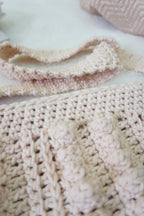 A close-up of a handmade crochet bag in a soft cream-beige color, showing textured bubble stitch details, with a matching strap.