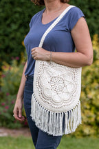 A woman wearing a blue top and jeans carries a large off-white crocheted shoulder bag with a long strap and fringe, standing in a garden with green foliage and pink flowers in the background.